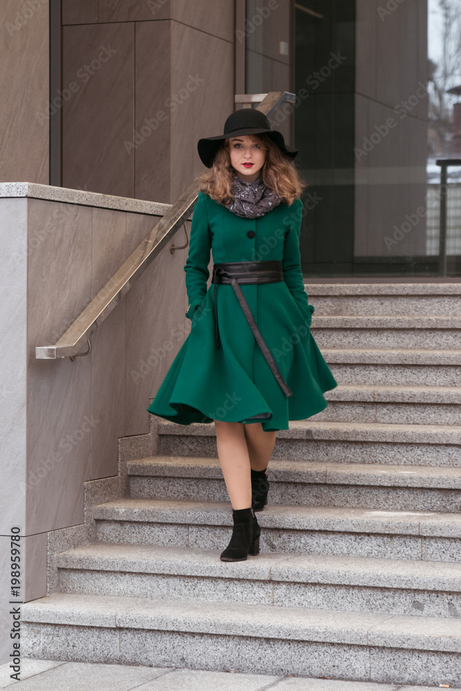 a girl in a green coat and a black hat walks down the stairs