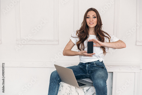 Beautiful girl in casual clothes sits on a chair and hold smaptphone with black screen in her hands. Freelancer at work demonstrates the phone. Girl with computer smiling photo