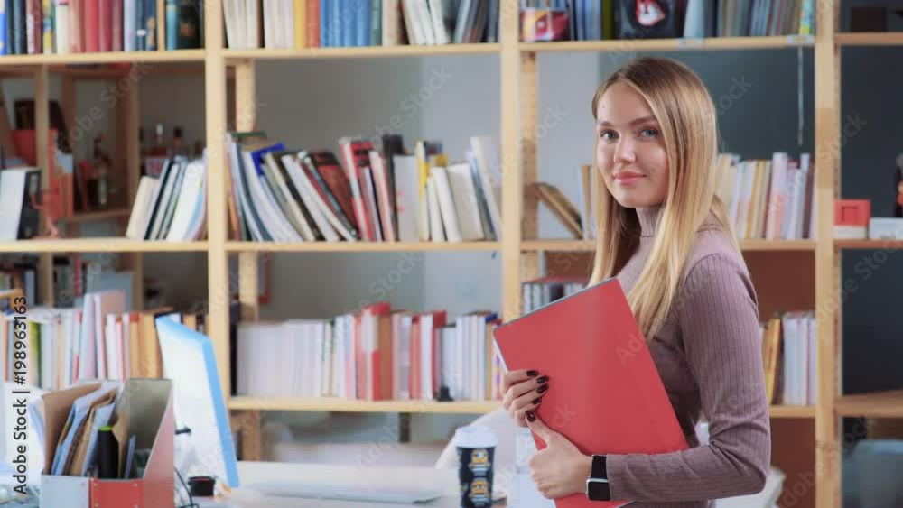 Portrait of a young and beautiful blonde with a red folder, large bookshelf on background. She looks at camera and smiles. Beautiful girl is an office employee, creative marketing agency. Secretary Portrait of a young and beautiful blonde with a red folder, large bookshelf on background. She looks at camera and smiles. Beautiful girl is an office employee, creative marketing agency. Secretary
