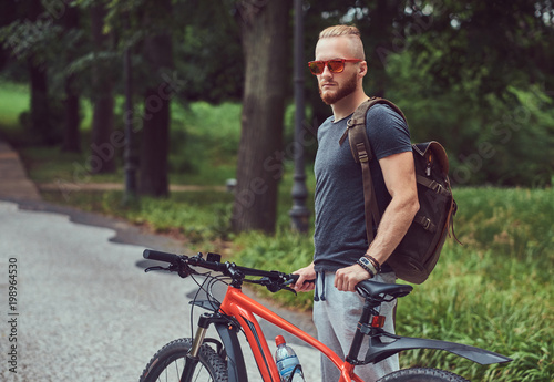 Fototapeta Naklejka Na Ścianę i Meble -  A handsome redhead male with a stylish haircut and beard dressed in sportswear and sunglasses walks in the park with a bicycle and backpack.