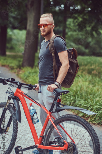 A handsome redhead male with a stylish haircut and beard dressed in sportswear and sunglasses walks in the park with a bicycle and backpack.