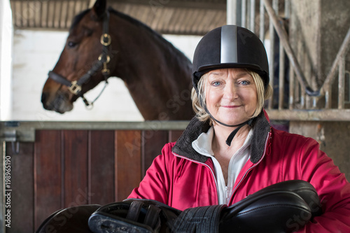 Portrait Of Mature Female Owner Holding Saddle In Stable With Horse