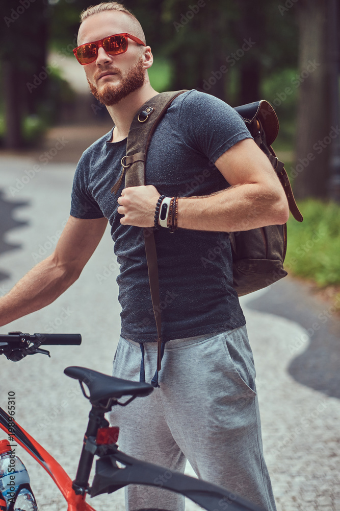 Obraz premium Przystojny rudy mężczyzna ze stylową fryzurą i brodą ubrany w sportową odzież i okulary przeciwsłoneczne spaceruje po parku z rowerem i plecakiem.