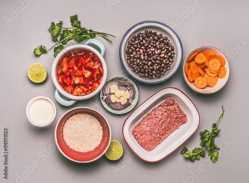 Flat lay of bowls with cooking ingredients for balanced one pan meal with beans, minced meat, rice and various cut vegetables on gray background, top view