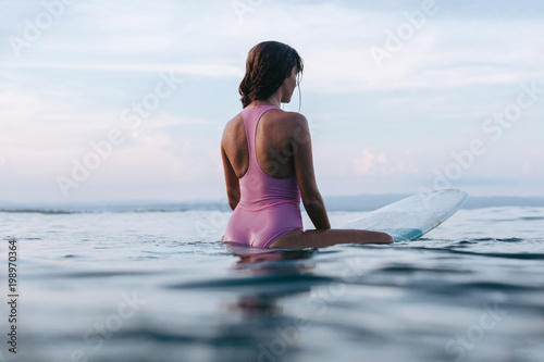 Photography back view of young sportswoman sitting on surfboard in water
