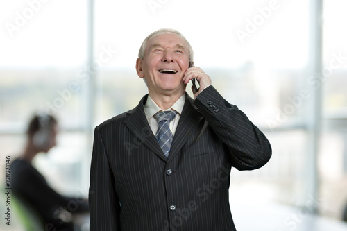 Happy cheerful businessman in suit talking on phone. Bright office background. © DenisProduction.com