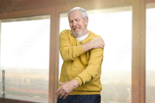 Old senior man with shoulder pain. Grandpa with shoulder inhury at home. Windows background.
