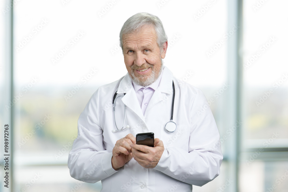 Old doctor standing, holding and typing on mobile phone. Smiling senior physician with phone looking at camera. Old doctor standing, holding and typing on mobile phone. Smiling senior physician with phone looking at camera.