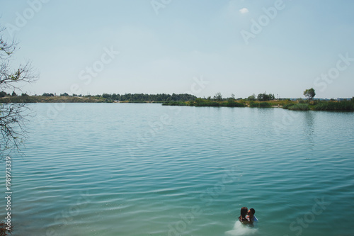 Wedding couple is hugging in azure blue lake splashes of water. Beautiful bride in puffy dress and groom are having fun. Summer passion crazy emotions photo on the seaside. Wet wedding clothes.