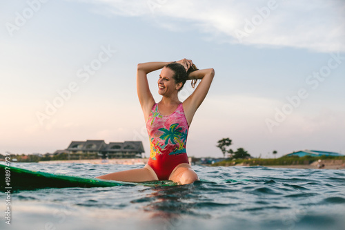 portrait of smiling woman readjusting hair while sitting on surfing board in ocean