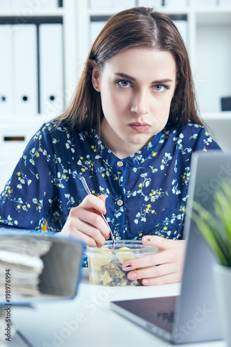 Business woman eating lunch at her workplace looking at the laptop screen. Folders with documents in the foreground. Dedline concept. photo