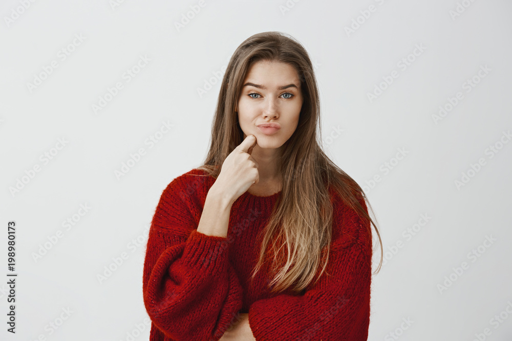 Studio shot of disappointed caucasian woman in red loose sweater, holding finger on chin and pouting, looking displeased and doubtful, seeing something uninteresting over gray background Studio shot of disappointed caucasian woman in red loose sweater, holding finger on chin and pouting, looking displeased and doubtful, seeing something uninteresting over gray background