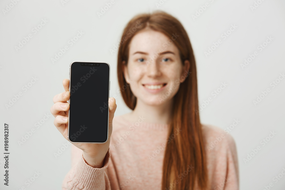 close-up portrait of redhead european woman advertising smartphone, smiling broadly, being blurred while focus is on gadget, standing over gray background. Model promotes new popular device close-up portrait of redhead european woman advertising smartphone, smiling broadly, being blurred while focus is on gadget, standing over gray background. Model promotes new popular device
