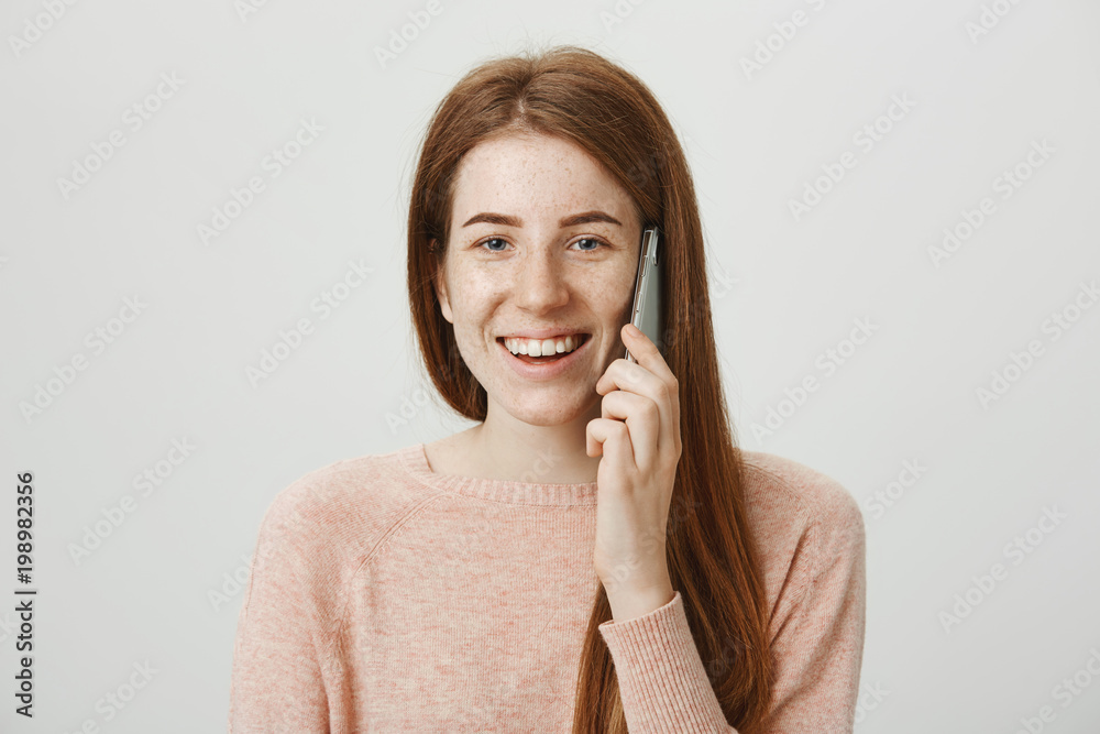 Φωτογραφία Studio portrait of beautiful caucasian redhead female model with freckles talking on smartphone while smiling and being in good mood, standing against gray background while advertising device
