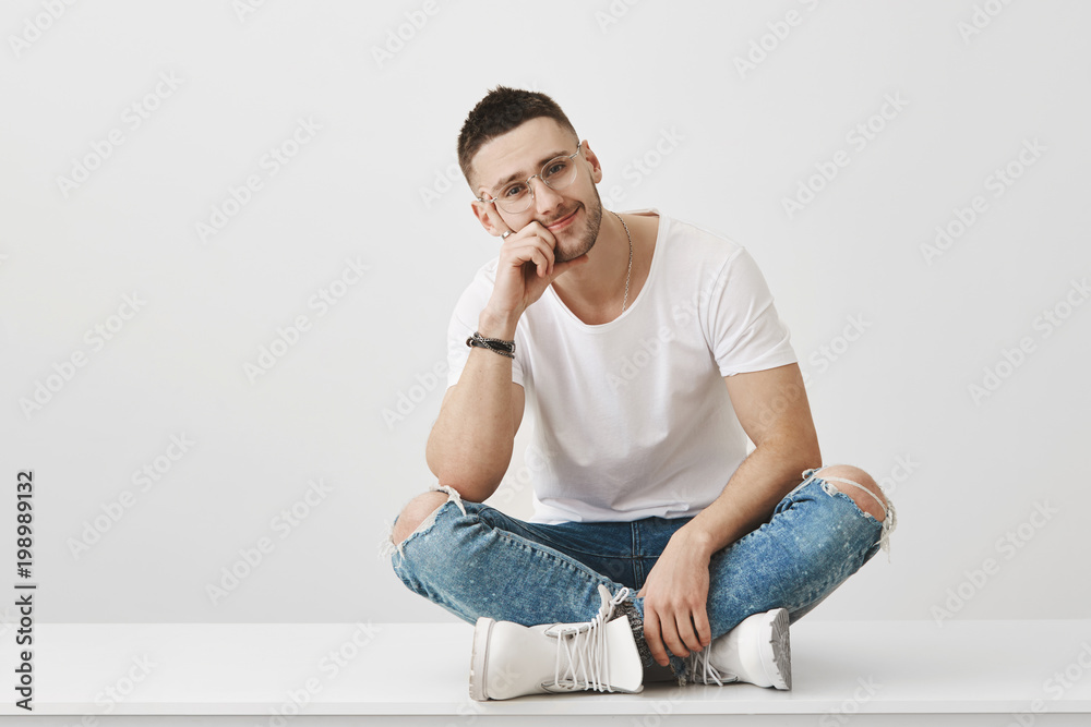 You are so boring. Studio shot of attractive caucasian unshaven guy in glasses, sitting with crossed legs on floor leaning head on hand and looking tired at camera over gray background. You are so boring. Studio shot of attractive caucasian unshaven guy in glasses, sitting with crossed legs on floor leaning head on hand and looking tired at camera over gray background.