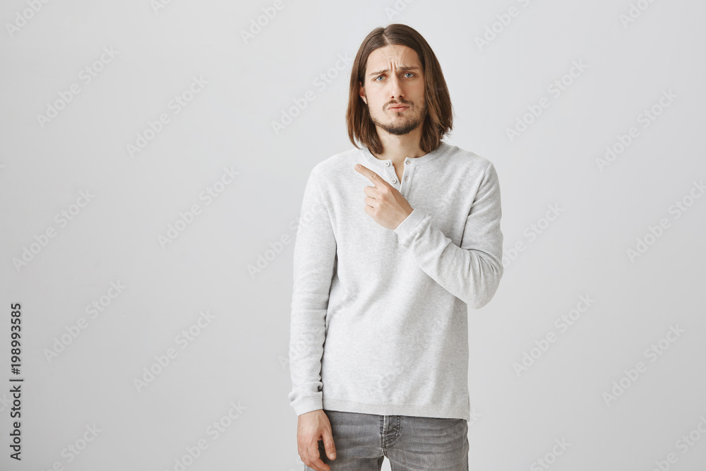 Man is very upset seeing sad commercial. Portrait of gloomy handsome guy with beard in casual white pullover pointing at upper left corner, frowning from sadness, standing over gray background