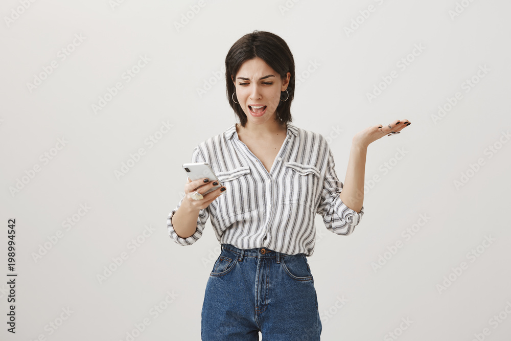 Woman received ridiculous message from unknown number. Annoyed and angry cute dark-haired businesswoman gesturing with raised palm while holding smartphone and typing answer, swearing from emotions