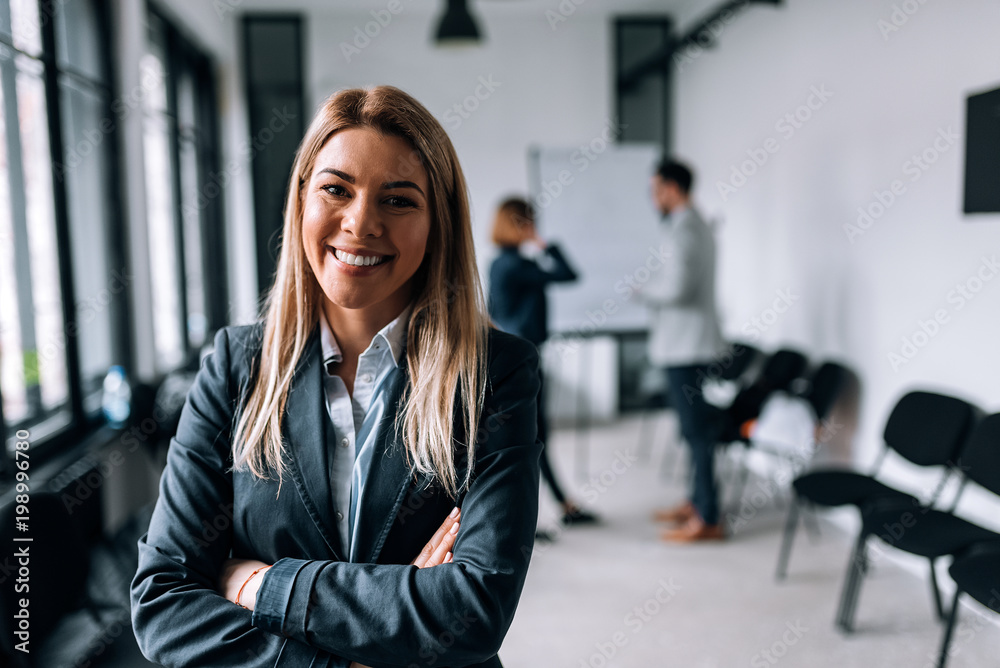 Portrait of a smiling blonde businesswoman with crossed arms, looking at camera. Collegues at the background. Portrait of a smiling blonde businesswoman with crossed arms, looking at camera. Collegues at the background.