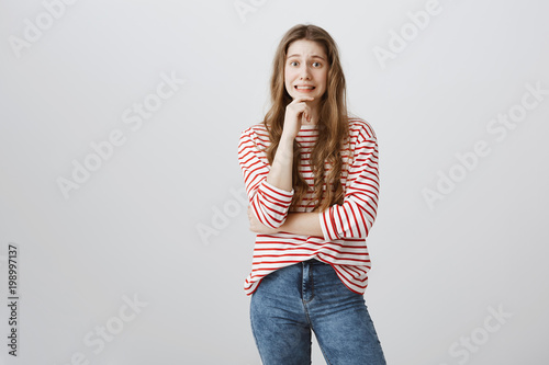 Yikes, mom saw broken vase. Studio shot of pretty embarrassed teenage girl holding hand on chin and grimacing with awkward expression, being guilty and hoping that nobody will know about her fault photo