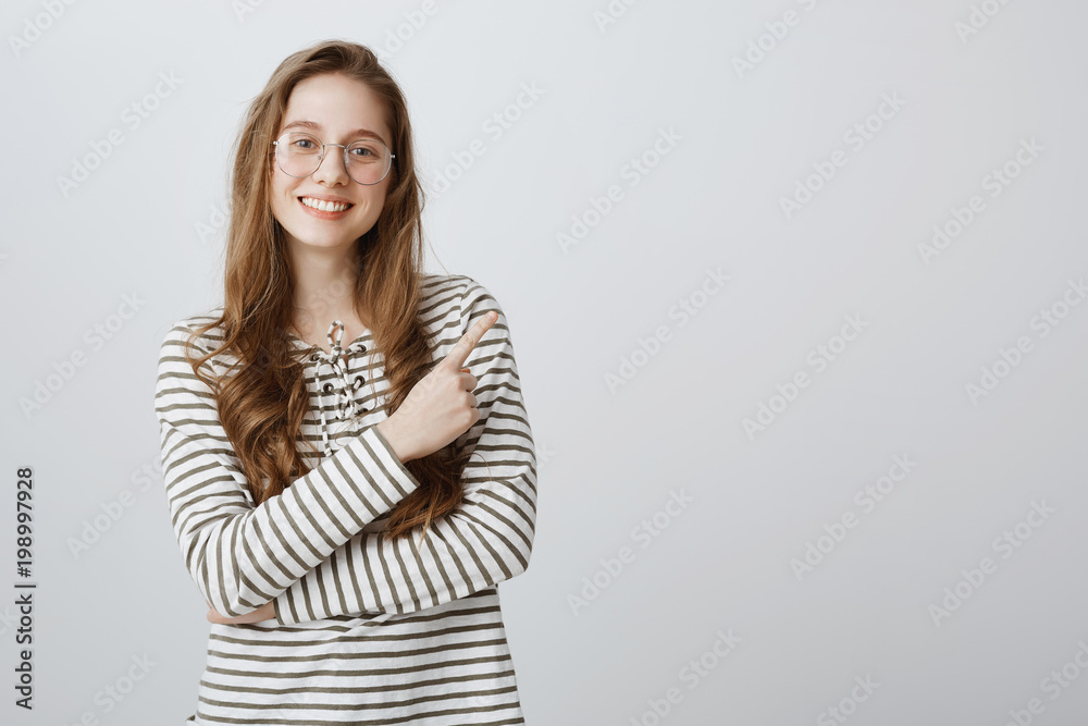 Shall we look at that copy space. Portrait of cheerful bright european girl in glasses pointing at upper right corner with index finger, smiling broadly, showing stand with goods while working in shop Shall we look at that copy space. Portrait of cheerful bright european girl in glasses pointing at upper right corner with index finger, smiling broadly, showing stand with goods while working in shop