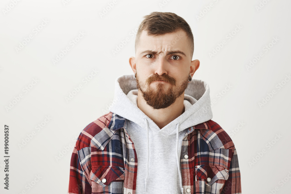 What are you looking at. Portrait of handsome stylish european student with flesh tunnels in ears, making serious and cool face while standing in trendy clothes over gray background, frowning