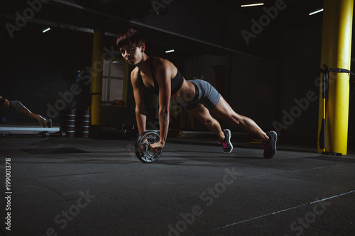 fitness girl doing exercises in gym with wheel in dark hall, sporty woman sexy in pose doing functional training, poster banner healthy lifestyle