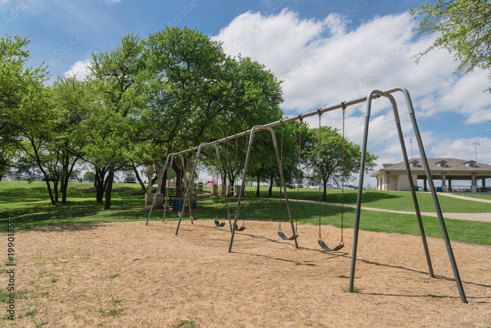 Close-up swing set 6 seats in nature park and tree lush at Ennis, Texas, USA. Outdoor sand playground hanging seat suspended from bar swings back and forth surround by leaves green, cloud blue sky