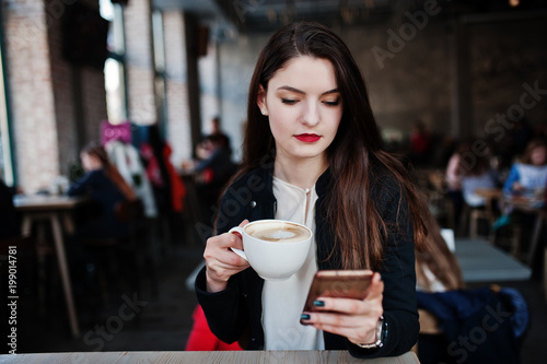 Brunette girl sitting on cafe with cup of cappuccino and looking at mobile phone.
