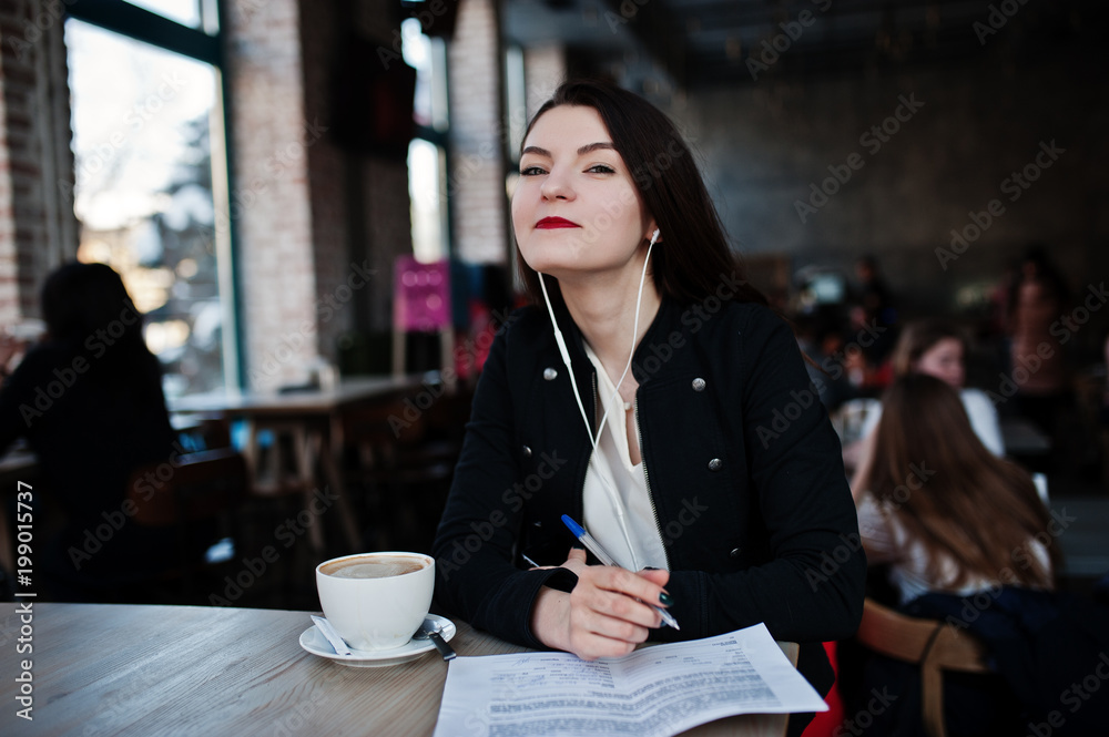 Brunette girl sitting on cafe with cup of cappuccino, listening music on headphones and write some documents. Brunette girl sitting on cafe with cup of cappuccino, listening music on headphones and write some documents.