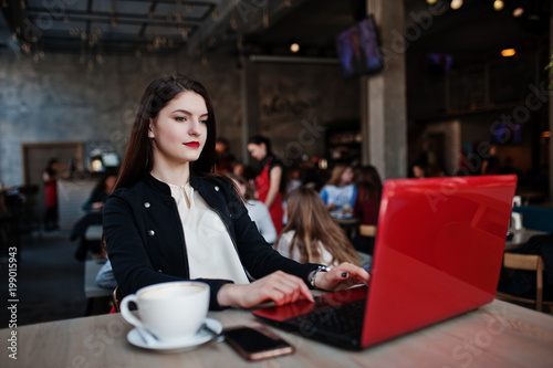 Brunette girl sitting on cafe with cup of cappuccino, working with red laptop.