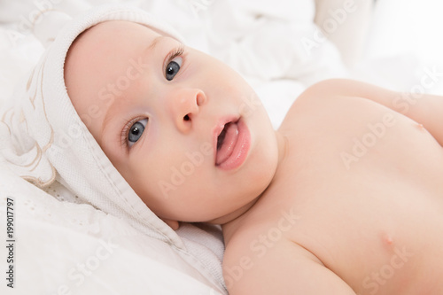 Happy baby boy wearing hooded towel lying on a bed after bath or shower. Young child with blue eyes looking at camera.