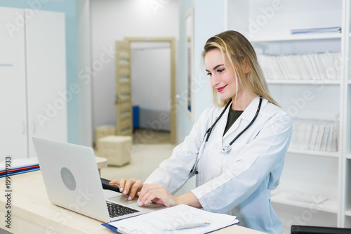 Smiling female physician using computer in her office in hospital. Young attractive Caucasian woman doctor therapist wearing medical uniform sitting at the desk