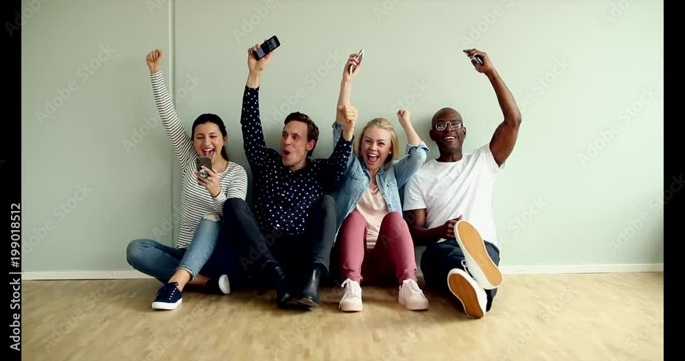 Diverse group of young designers using their cellphones and cheering together while sitting on a floor during a break in a modern office Diverse group of young designers using their cellphones and cheering together while sitting on a floor during a break in a modern office