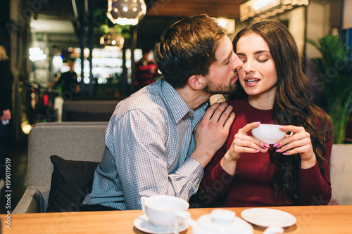 Attractive man is sitting with his girlfrien at the table and kissing her. She is holding a cup of tea and receiving a kiss. Lovely picture of lovely couple.
