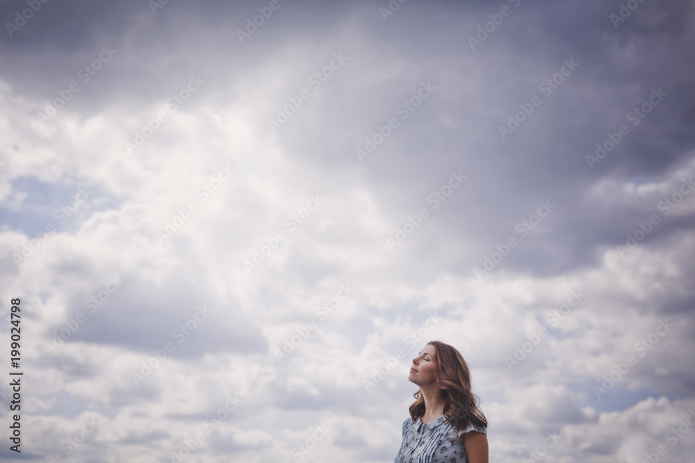A beautiful young brunette woman with long hair in a blue dress stands with her eyes closed against the blue clouds on a warm summer day. A beautiful young brunette woman with long hair in a blue dress stands with her eyes closed against the blue clouds on a warm summer day.
