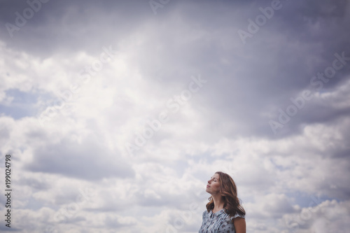 A beautiful young brunette woman with long hair in a blue dress stands with her eyes closed against the blue clouds on a warm summer day.