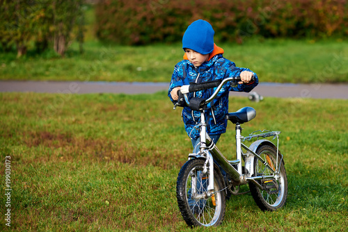 Cute european boy is standing near his bicycle in the autumn city park.
