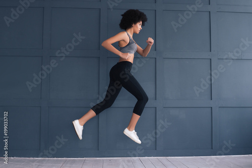 Keep in shape for your benefit! Gorgeous good-looking young athlete girl running in the air and wearing tight sport clothes.