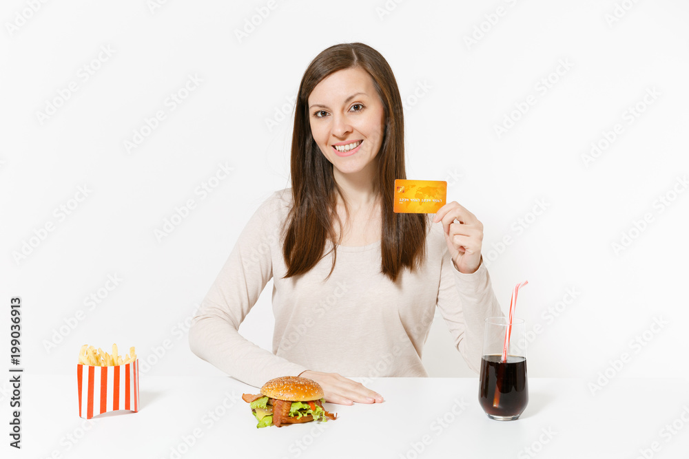 Happy woman sitting at table with credit card, burger, french fries, cola in glass bottle isolated on white background. Proper nutrition or American classic fast food. Advertising area with copy space Happy woman sitting at table with credit card, burger, french fries, cola in glass bottle isolated on white background. Proper nutrition or American classic fast food. Advertising area with copy space