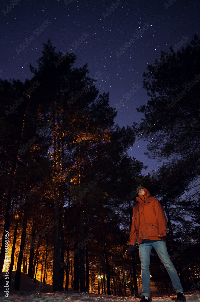 A man in an orange jacket and jeans looks at the starry sky in a pine forest in winter..