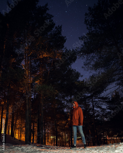 A man in an orange jacket and jeans looks at the starry sky in a pine forest in winter..