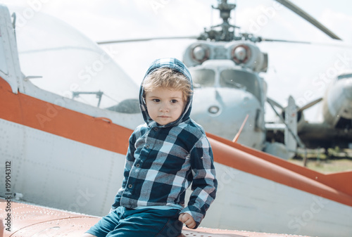Cute boy sits on wing of old plane in aviation museum. Textured grunge old helicopter and sky with clouds on background. Kid on excursion to museum of aviation in open air. Air forces history concept.