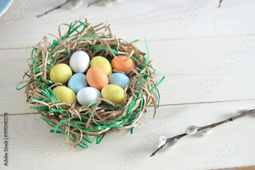 A nest of strips of paper, imitating a bird's nest. In the nest lie candy in the form of eggs. In the frame there is a willow branch. Light background. View from above. Close-up.