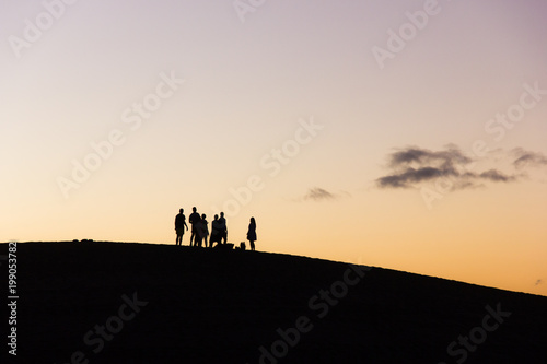 Group of friends enjoying nice sunset on top of Maspalomas sand dunes in Canary Islands. Adventure explorers walking on a different planet concept