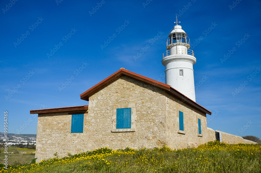 White lighthouse on blue sky background. Summer, sea and hope concept. Stone building with red roof tiles and blue windows. Green field with yellow flowers. Cyprus, Paphos.