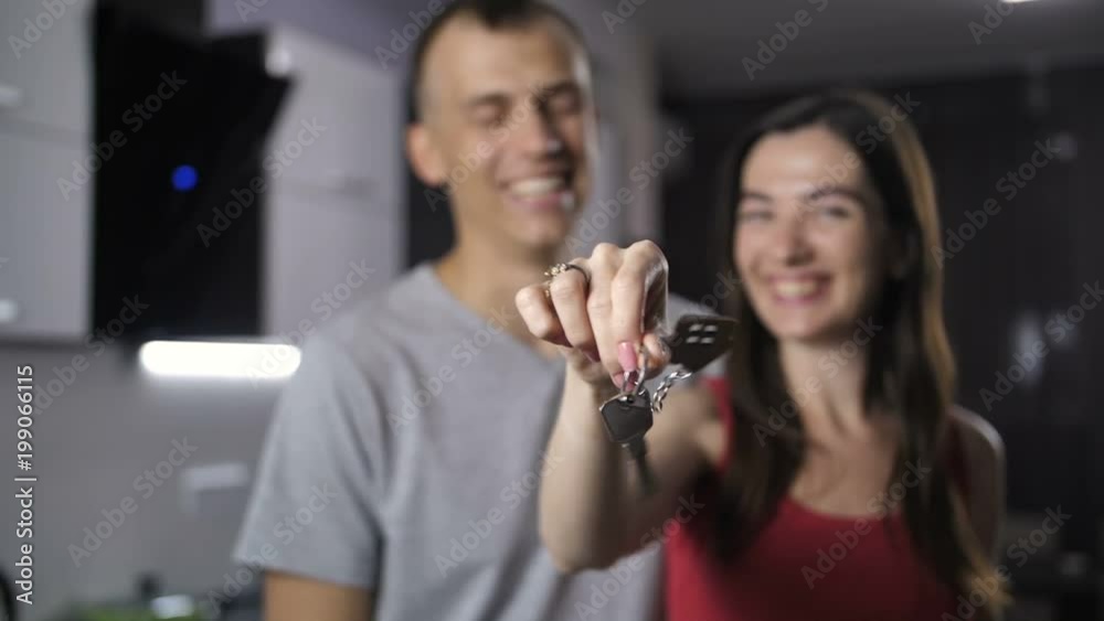 Foreground keys with wooden house shaped key ring, background happy smiling couple just purchased real estate. Excited male kissing beloved woman's head as they are holding apartment keys. Slow motion Foreground keys with wooden house shaped key ring, background happy smiling couple just purchased real estate. Excited male kissing beloved woman's head as they are holding apartment keys. Slow motion