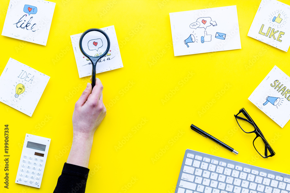 Socail media icons on work desk of marketing expert. Digital promotion of goods and services. Yellow background top view copy space. Hand with magnifier