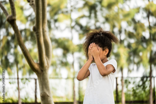 A Little girl is standing in a park, covering her face with her hands