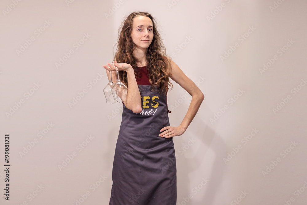Cheerful girl poses in front of the camera in gray apron. Young cook holding two glasses in her hand and smiles. Cheerful girl poses in front of the camera in gray apron. Young cook holding two glasses in her hand and smiles.