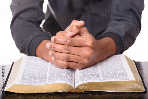 Hands of a young man folded praying over a Bible, hands over soft focus Bible